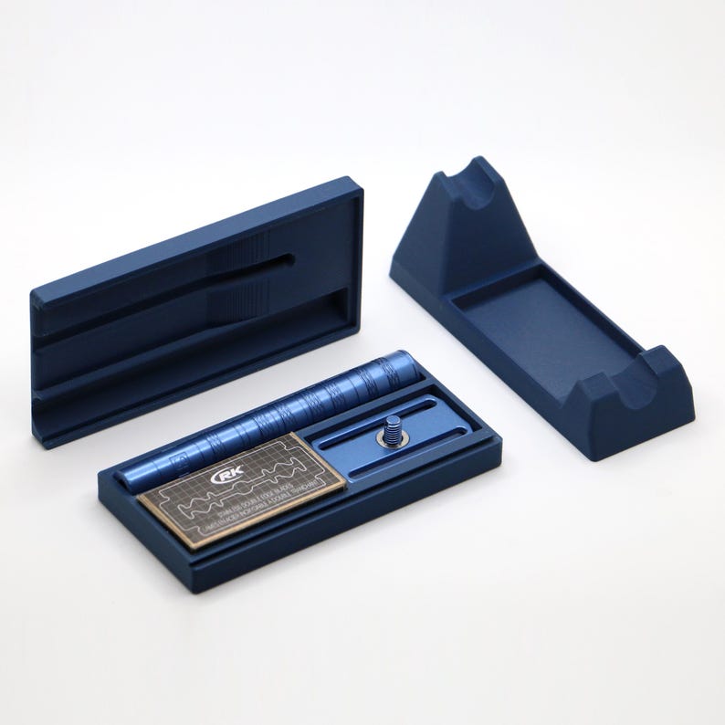 K&ouml;nnte beinhalten: Ein mattes marineblaues Rasierhobel-Set bestehend aus einem zerlegten Griff, einem Rasierkopf mit Gewindestift, einem rechteckigen Aufbewahrungsetui mit Deckel und einem St&auml;nder. Eine kleine Metallklingenpackung mit der Aufschrift RK liegt im Etui.