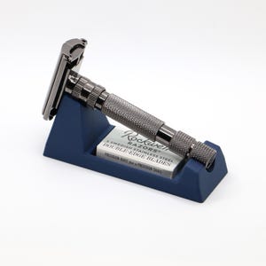 May include: A dark gray safety razor with a textured handle rests on a dark blue stand. A box of double-edge razor blades is in the stand. The text on the box reads "Rockwell Razors" and "Precision Built for a Precision Shave."