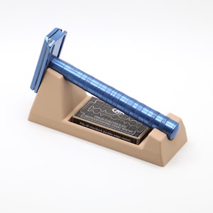 May include: A metallic blue double edge safety razor resting on a tan stand. A small black box of stainless double edge blades is tucked into the stand, featuring the text "Fits all standard double edge razors" and "STAINLESS DOUBLE EDGE BLADES".