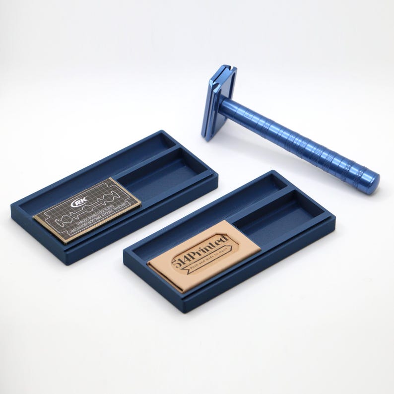 K&ouml;nnte beinhalten: Ein metallisch blauer Sicherheitsrasierer mit strukturiertem Griff und zwei rechteckigen blauen Ablageschalen. Eine Schale enth&auml;lt eine schwarze Klingenverpackung mit der Aufschrift RK, die andere eine beige Verpackung mit dem Text 514Printed.