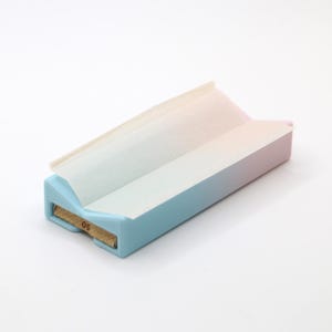 May include: A light blue plastic rolling machine with a gradient pink and white paper sheet extending from the top. The device features a small brown textured roller visible at the front end with the number 50 printed on its side surface.