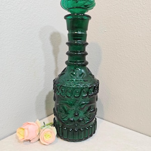 May include: A vintage green glass decanter with a decorative stopper. The bottle features embossed floral and scrollwork patterns. The decanter is approximately 12 inches tall. Two pink and yellow roses are in the foreground.