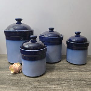 May include: Four blue ceramic jars with lids. The jars are in varying sizes, with a dark blue top and a lighter blue body. A pink rose is in the foreground. These jars are suitable for kitchen storage.
