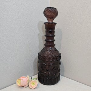 May include: A dark purple glass decanter with a decorative stopper. The bottle has an ornate, embossed design and is approximately 12 inches tall. Two small pink and yellow roses are in the foreground.