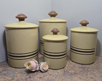 May include: Four matching beige ceramic canisters with lids and dark brown stripes. The canisters have wooden knobs on the lids. Two pink roses are in front of the canisters. The canisters are on a brown and gray surface.