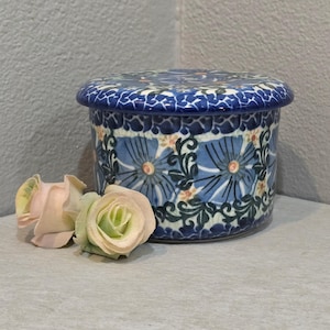 May include: A round, blue and white ceramic butter dish with a floral design. The lid and body feature a repeating pattern of blue flowers and green leaves. Two artificial roses in shades of pink and green are next to the dish.
