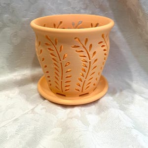 May include: A peach-colored ceramic planter with a matching saucer. The planter features cut-out leaf designs around the sides and top. The pot is designed to hold plants and flowers. The planter is sitting on a white surface.
