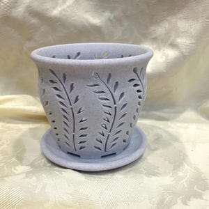 May include: A light lavender speckled plant pot with a matching saucer. The pot features a repeating cut-out leaf pattern around the exterior, creating a series of vertical fern-like designs that allow for ventilation and visibility through the walls.