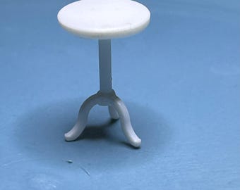 May include: A small, white, round table with a single stem and three curved legs. The table is made of a solid material and is designed for decorative purposes. The table is approximately 2 inches in height.