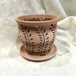 May include: A terracotta-colored ceramic planter with a matching saucer. The planter features cut-out leaf designs around the sides and a decorative rim. The planter is designed for indoor or outdoor use.