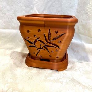 May include: A square, terracotta-colored planter with a matching saucer. The planter features a bamboo leaf design and small flower cutouts. The planter is designed for indoor or outdoor use.