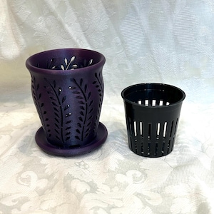 May include: A dark purple planter with a cutout leaf pattern and a matching saucer, accompanied by a separate black plastic slotted inner pot. The planter features a flared rim and a cylindrical shape, while the inner pot has vertical slits.