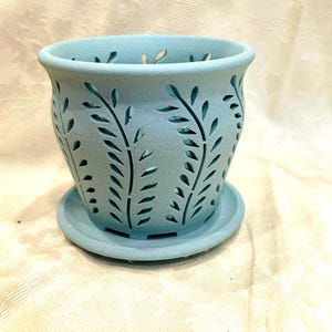 May include: A light blue ceramic planter with a matching saucer. The planter features a cut-out leaf design around the sides. The planter is approximately 5 inches tall and 5 inches wide.