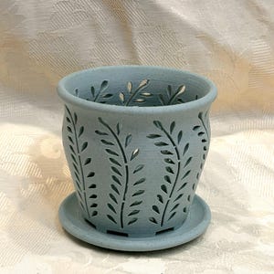 May include: A light blue speckled ceramic planter with a matching saucer. The cylindrical pot features a repeating cut-out pattern of vertical fern-like leaves around the exterior, allowing light to pass through the openings in the vessel walls.