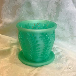 May include: A green glass planter with a matching saucer. The planter has a detailed leaf pattern and a slightly flared rim. The color is a vibrant, translucent green, and the design is reminiscent of vintage glassware.