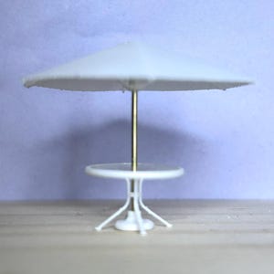 May include: Miniature white patio set with a round table and umbrella. The umbrella has a white canopy and a gold pole. The table has a white top and a three-legged base. The set is designed for a dollhouse or miniature scene.