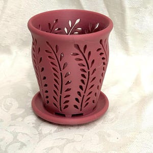 May include: A dusty rose-colored cylindrical planter featuring a decorative cut-out pattern of vertical fern-like leaves. The planter includes a matching circular saucer base, providing a cohesive look for this functional plant container.
