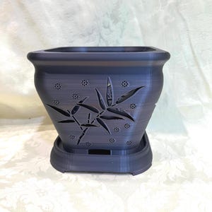 May include: A dark gray square-shaped planter with a bamboo leaf design. The planter has a matching saucer and decorative cutouts. The planter is made of a solid material and is suitable for indoor or outdoor use.