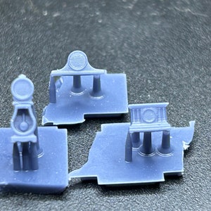 May include: Three 3D printed plastic models of a clock tower with a circular clock face and three pillars. The models are light blue and are on a gray surface.
