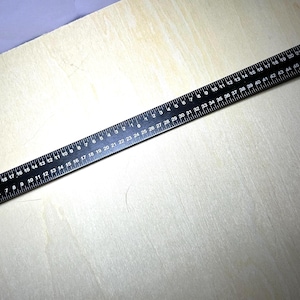 May include: A black metal ruler with white markings, measuring up to 50 inches. The ruler has both inch and HO scale measurements. The ruler is lying on a light-colored wooden surface.