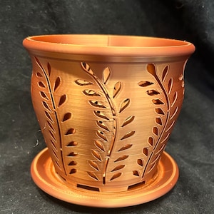 May include: A copper-colored planter with a decorative leaf design. The planter has drainage holes and a matching saucer.