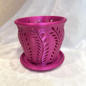 May include: A magenta-colored, 3D-printed plant pot with a matching saucer. The pot features a decorative cut-out leaf design around the sides. The pot is designed to hold small plants or flowers. The pot measures approximately 5 inches in diameter and 4 inches tall.