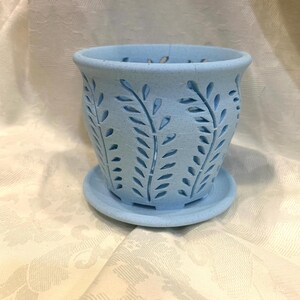 May include: A light blue ceramic planter with a matching saucer. The planter features cut-out leaf designs around the sides. The pot is designed to hold plants and flowers. The planter is approximately 5 inches in diameter and 4 inches tall.