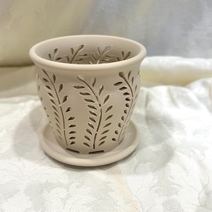 May include: A matte beige ceramic planter with a matching saucer, featuring a repeating cut-out leaf pattern around the exterior. The cylindrical vessel has a smooth, flared rim and a series of vertical, stylized foliage designs.