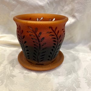 May include: A ceramic plant pot with a matching saucer, featuring a gradient color scheme that transitions from orange at the top to dark brown at the base. The exterior is decorated with vertical cut-out patterns resembling fern leaves.