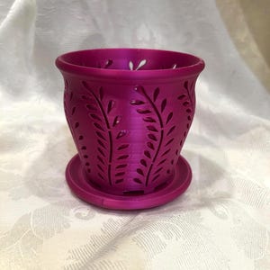 May include: A magenta-colored plant pot with a matching saucer. The pot features a decorative leaf pattern cut into the sides. The pot is designed to hold small plants or flowers. The pot is made of a hard plastic material.