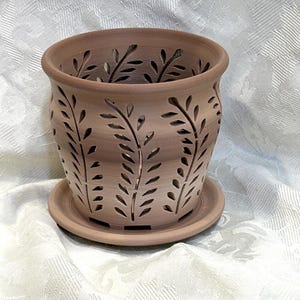 May include: A brown ceramic planter with a matching saucer. The planter features cut-out leaf designs around the sides. The pot is designed to hold plants and flowers. The planter is approximately 6 inches in diameter and 5 inches tall.