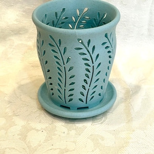May include: A light blue speckled plant pot with a matching saucer. The cylindrical pot features a repeating cut-out pattern of vertical leafy branches around its exterior, allowing light to pass through the openings in the ceramic-like material.