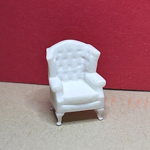 May include: A miniature white armchair with a tufted back and rolled arms. The small chair has four delicate legs and is likely a dollhouse or model furniture piece. The chair is set against a red background.