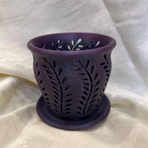 May include: A dark purple ceramic planter with a decorative leaf pattern. The planter has a drainage hole and a matching saucer.