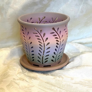 May include: A decorative ceramic planter with a matching saucer. The planter is cylindrical with cut-out leaf designs and a gradient of colors, transitioning from lavender to green. The planter is designed for indoor or outdoor use.