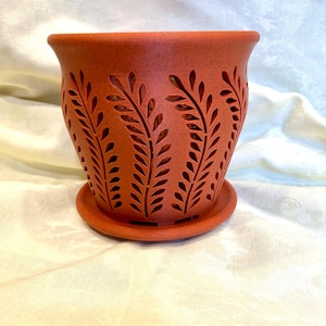 May include: A terracotta-colored ceramic planter with a matching saucer. The planter features a series of cut-out leaf designs around its exterior. The pot has a slightly flared rim and a round base, suitable for indoor or outdoor use.