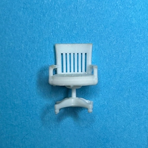 May include: A small, white miniature office chair with a vertical slat back, armrests, and a swivel base. The chair is set against a solid blue background, showcasing its design and details.