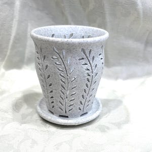 May include: A light gray speckled ceramic planter with a cylindrical shape and a matching circular saucer. The exterior features vertical cutouts in a repeating leaf or fern pattern that extend from the top rim down to the base of the pot.