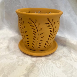 May include: A yellow-orange ceramic flower pot with a matching saucer. The pot features cut-out leaf designs around the sides and small drainage holes near the rim. The pot is designed for indoor or outdoor use.