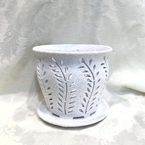 May include: A white ceramic planter with a matching saucer. The planter has a cylindrical shape with cut-out leaf designs. The surface has a speckled texture. The planter is designed for indoor or outdoor use.