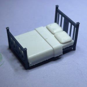 May include: A miniature bed with a dark blue frame and a white mattress, pillows, and bedding. The bed frame has a slatted headboard and footboard. The bed is a small scale model, likely for a dollhouse or miniature scene.