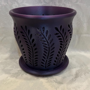 May include: A deep purple ceramic planter with a matching saucer. The planter features a series of leaf-shaped cutouts around the exterior. The pot has a wide opening and a slightly flared rim. The planter is designed for indoor or outdoor use.