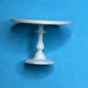May include: A white miniature table with a round top and a pedestal base. The table is made of plastic and is about 3 inches in diameter.