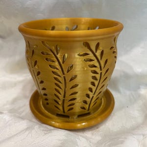 May include: A golden-colored ceramic planter with a matching saucer. The planter features cut-out leaf designs around the sides. The rim of the planter is rounded. The planter is designed to hold plants.