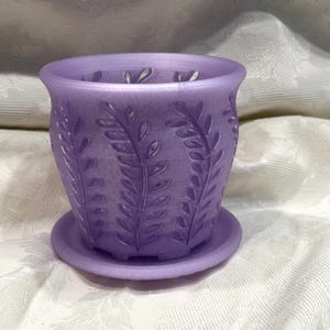May include: A lavender-colored plant pot with a matching saucer. The pot features a detailed leaf pattern around the exterior. The pot is designed to hold small plants and flowers.