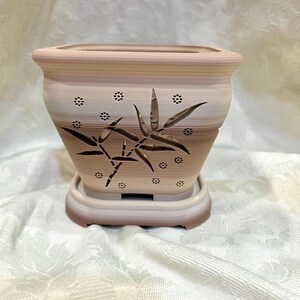 May include: A square, beige ceramic planter with a bamboo leaf design. The planter has a matching saucer and small decorative holes. The bamboo design is brown and the planter has a striped pattern.