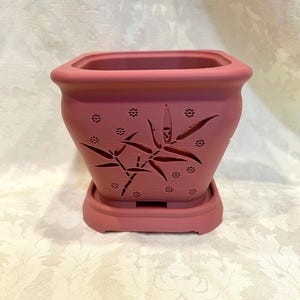 May include: A dusty rose-colored square ceramic planter with a matching saucer. The planter features cut-out bamboo and floral designs. The planter is designed for indoor or outdoor use.