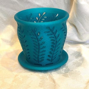 May include: A teal-colored ceramic planter with a matching saucer. The planter features a cut-out leaf design around the sides and top. The pot is round with a slightly flared rim. The saucer is a simple, flat circle.