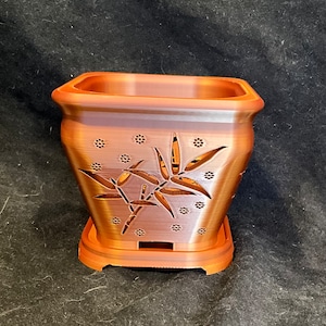 May include: A square terracotta-colored planter with a decorative cutout design of bamboo leaves and small circles. The planter has a matching base.