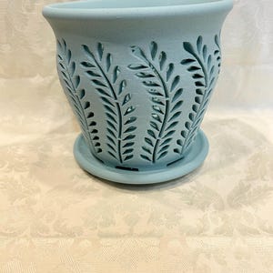 May include: A light blue ceramic planter with a matching saucer. The planter features a cut-out leaf design around the sides. The pot is approximately 6 inches in diameter and 5 inches tall, perfect for indoor or outdoor plants.